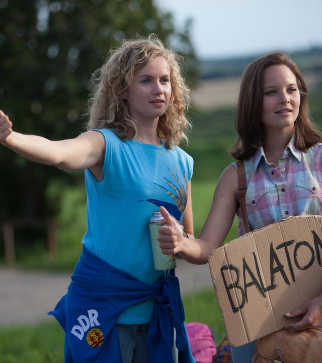 "Honigfrauen - 1. Urlaub im Paradies": Catrin Streesemann (Cornelia Gröschel) und Maja Streesemann (Sonja Gerhardt), die ein Pappschild mit der Aufschrift "Balaton" in der Hand hält, heben ihren Daumen, um per Anhalter mitgenommen zu werden.