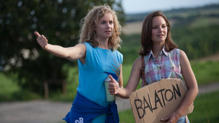"Honigfrauen - 1. Urlaub im Paradies": Catrin Streesemann (Cornelia Gröschel) und Maja Streesemann (Sonja Gerhardt), die ein Pappschild mit der Aufschrift "Balaton" in der Hand hält, heben ihren Daumen, um per Anhalter mitgenommen zu werden.