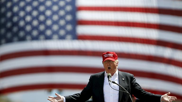 "Trumps Weg an die Macht": Donald Trump steht mit ausgebreiteten Armen vor einem Rednerpult mit Mikrofon. Im Hintergrund eine bildfüllende US-Flagge.