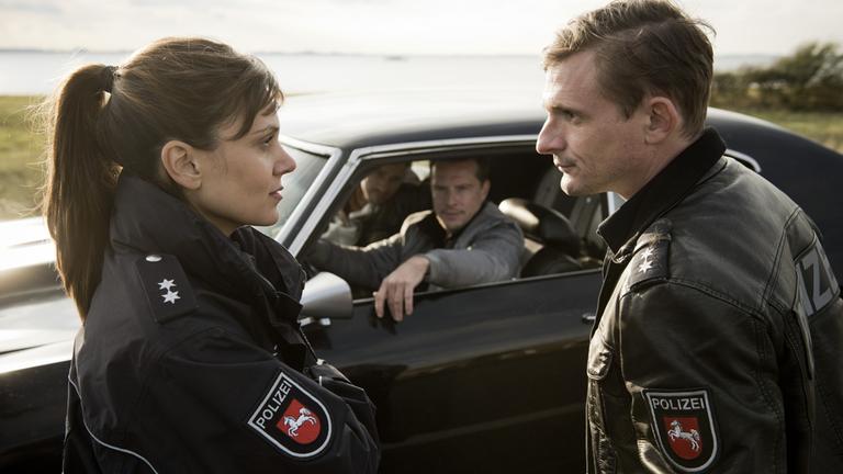 "Friesland - Krabbenkrieg": Süher Özlügül (Sophie Dal) und Jens Jensen (Florian Lukas) blicken sich ernst in die Augen. Sie stehen vor einem Auto, in dem Luke (Florian Panzner) am Steuer sitzt.