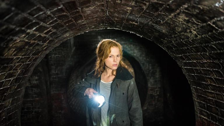"Im Tunnel": Maren (Maria Simon) befindet sich in einem dunklen, unterirdischen Tunnelsystem. Sie hält eine Taschenlampe in der Hand und scheint etwas entdeckt zu haben.