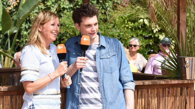 "ZDF-Fernsehgarten on tour": Andrea Kiewel und Lutz van der Horst, beide mit ZDF-Mikrophonen in der Hand, stehen lässig an einer Bude.