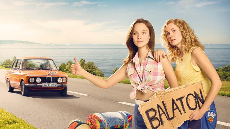 "Honigfrauen": Catrin Streesemann (Cornelia Gröschel) und Maja Streesemann (Sonja Gerhardt), die ein Pappschild mit der Aufschrift 'Balaton" in der Hand hält, heben ihren Daumen, um per Anhalter mitgenommen zu werden.