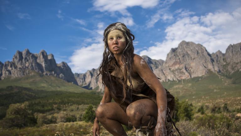 "Terra X: Die ersten Menschen - Aus Afrika in die ganze Welt": Eine Homo Erectus-Frau hockt vor einer Gebirgslandschaft.