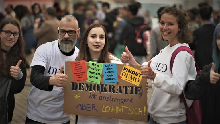 "Update für die Demokratie": Drei Personen halten ein Schild aus Pappe in den Händen. Auf dem steht: Demokratie ist anstrengend, aber lohnt sich!
