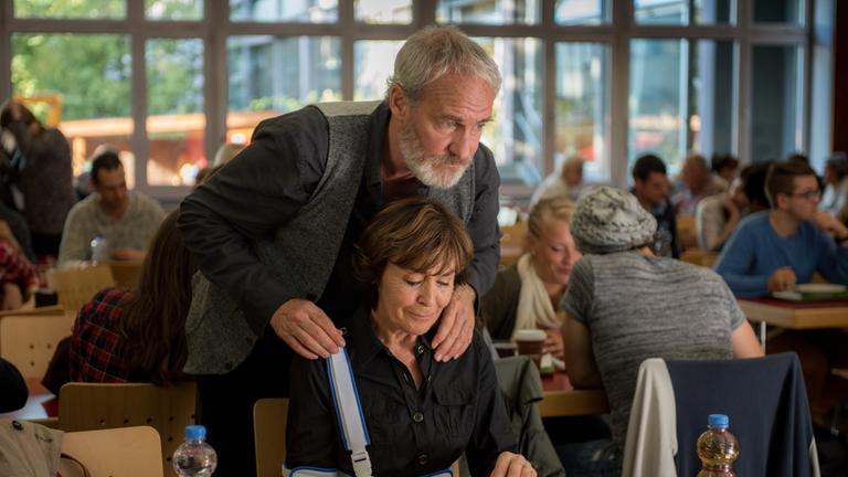 "Was ich von dir weiß": Ruth (Thekla Carola Wied) sitzt vor ihrem Essen. Georg (August Zirner) steht hinter ihr und hat die Hände auf ihren Schultern.