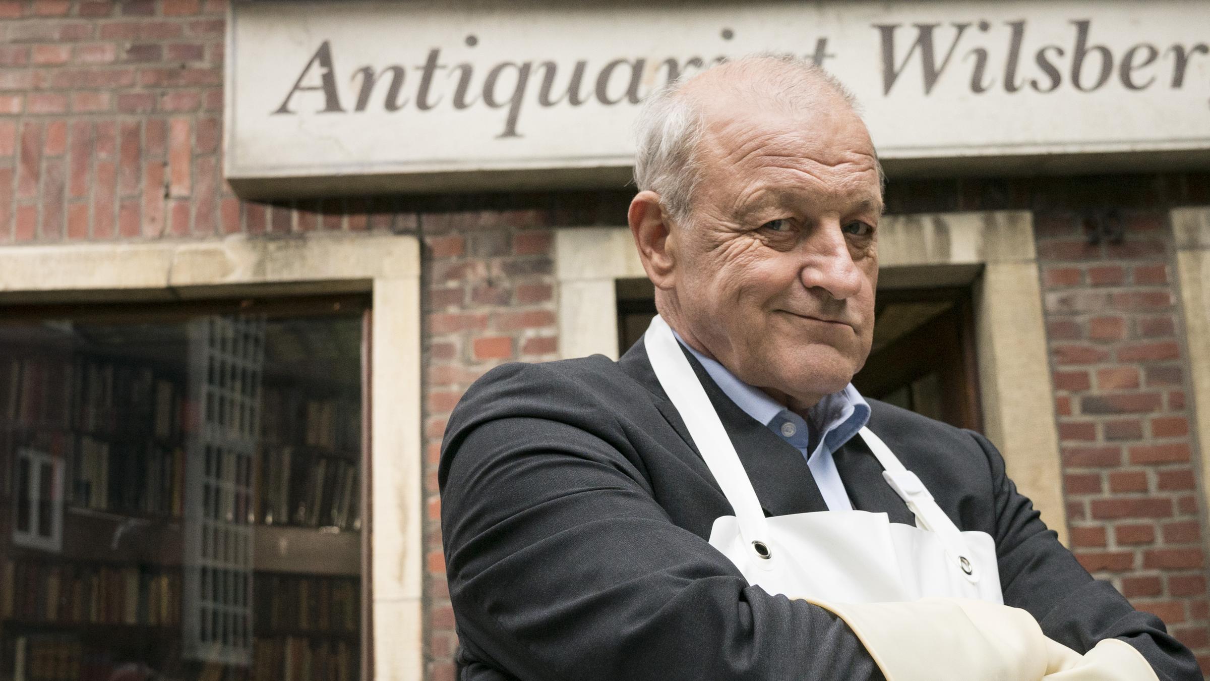 "Wilsberg - MünsterLeaks": Georg Wilsberg (Leonard Lansink) steht mit Schlachterschürze und -handschuhen vor seinem Antiquariat und schaut in die Kamera.