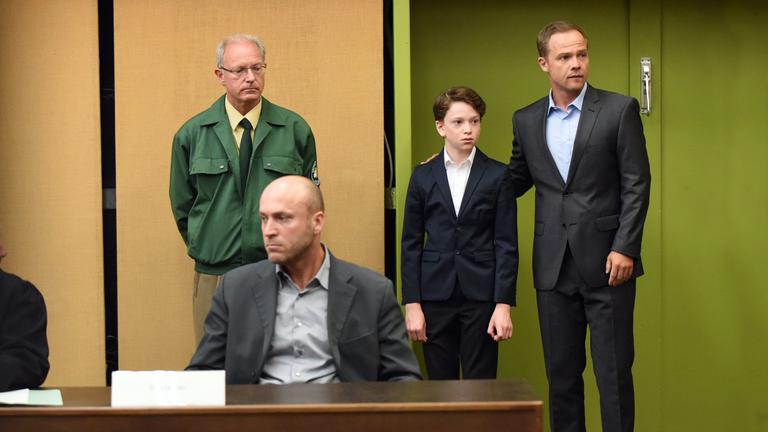 "Der Polizist, der Mord und das Kind": In einem Gerichtssaal sitzt Alexanders Vater (Ilir Rexhepi) am Tisch. Hinter ihm stehen Alexander (Joshio Marlon) und Opferschutzkommissar Carlos Benede (Matthias Koeberlin). Benede hat seinen rechten Arm um Alexanders Schulter gelegt.