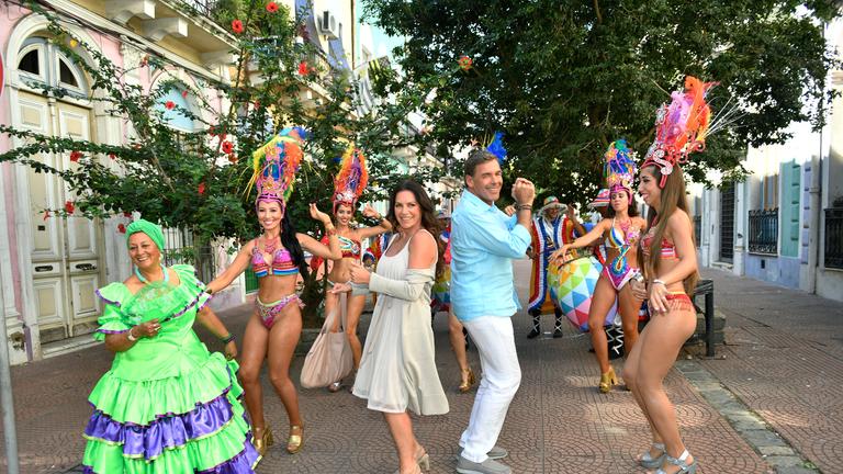 "Das Traumschiff: Uruguay": Kapitän Victor Burger (Sascha Hehn) und Caroline (Christine Neubauer) tanzen Rücken an Rücken auf einem Platz. Um sie herum tanzen leicht bekleidete Tänzerinnen, die alle einen großen Kopfschmuck tragen. Im Hintergrund steht ein Trommler.