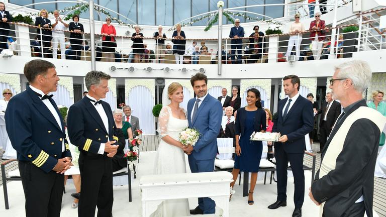  "Kreuzfahrt ins Glück - Hochzeitsreise nach Sardinien": Das Brautpaar Yvonne (Mira Bartuschek) und Paul (Raphael Vogt) steht an Deck des Schiffs vor einer Kirchenbank, hält sich an den Händen und blickt den Bordpfarrer (Jürgen Kuvik) an, der vor ihnen steht. Kapitän Burger (Sascha Hehn), Dr. Sander (Nick Wilder), Laura (Amy Mußul) und Tom (Jan Hartmann) stehen um beide herum versammelt.