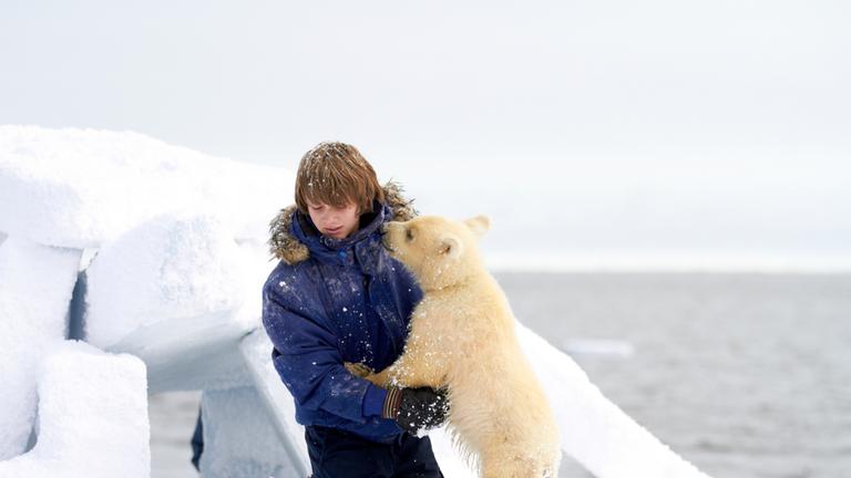"Midnight Sun - Eisbär auf Reisen": Luke (Dakota Goyo) und das Eisbären-Junge Pisu, sind auf einer Eisscholle gestrandet. Luke kniet vor dem kleinen Pisu, der auf den Hinterfüßen steht und mit seiner Schnauze Luke's Jackenkragen beschnüffelt.
