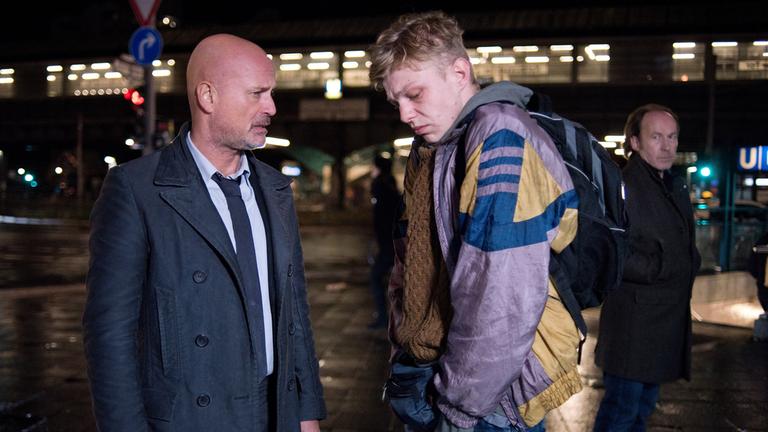 "Der Kriminalist – Die offene Tür": Schumann (Christian Berkel) steht mit Max (Leonard Kunz) nachts auf einer Berliner Straße. Der Kommissar sieht den Jungen mit ernstem Blick an. Max weicht seinem Blick aus und schaut zu Boden. Etwas entfernt von den beiden steht Ulli Grützke (Ulrich Noethen) und sieht zu ihnen herüber.
