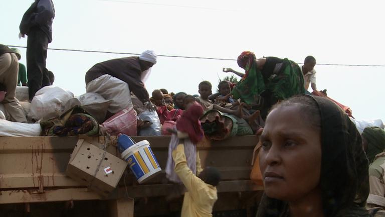 "Cahier africain": Frau steht vor einem mit Menschen und Gepäck beladenen LKW, auf den ein Mann ein kleines Kind hebt.