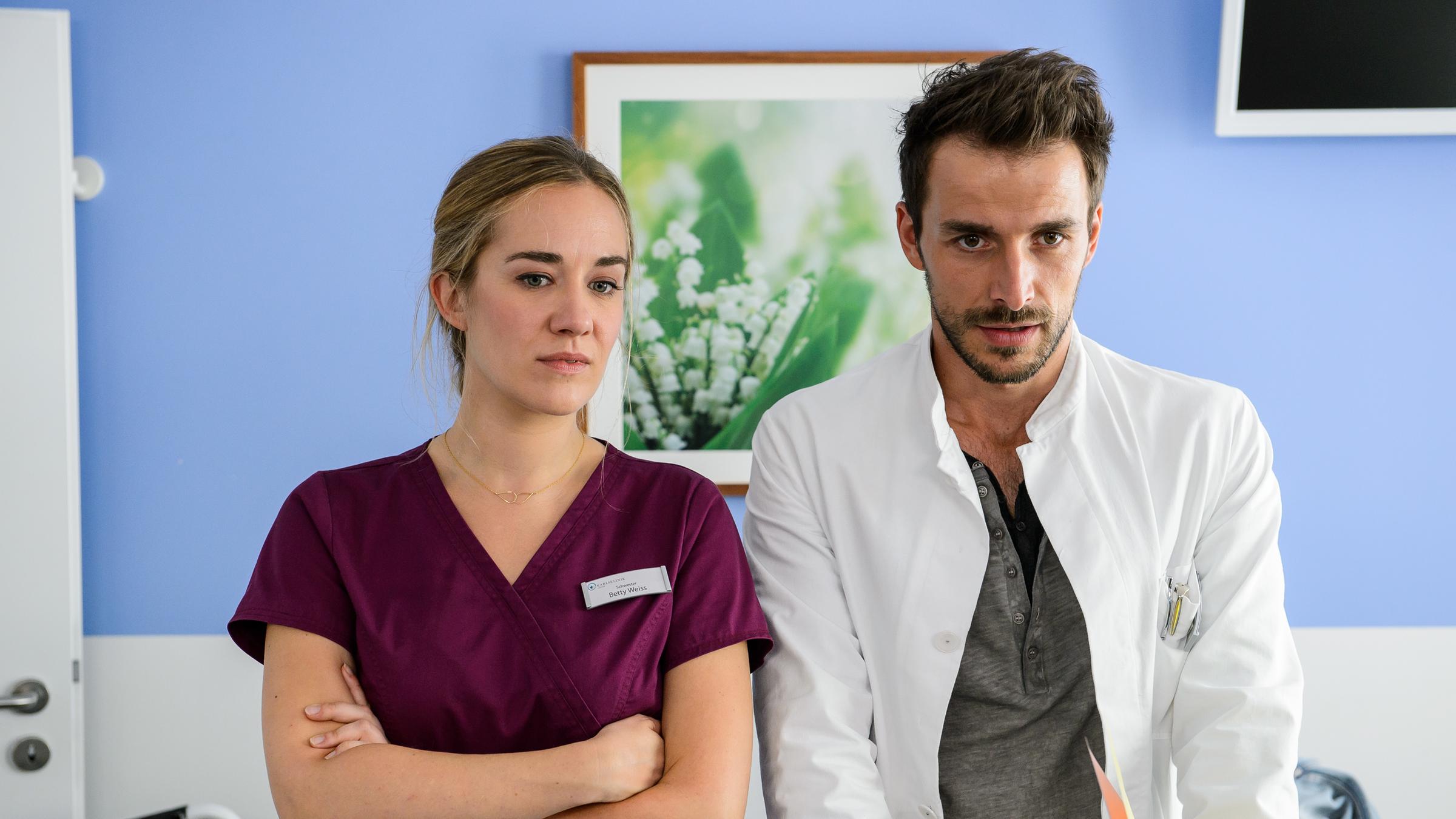 "Bettys Diagnose - Verletzte Gefühle": Betty Weiss (Annina Hellenthal) steht mit verschränkten Armen neben Dr. Frank Stern (Max Alberti) am Krankenbett eines Patienten. Beide schauen nach vorne.