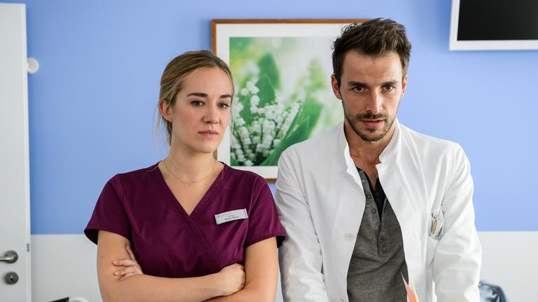 "Bettys Diagnose - Verletzte Gefühle": Betty Weiss (Annina Hellenthal) steht mit verschränkten Armen neben Dr. Frank Stern (Max Alberti) am Krankenbett eines Patienten. Beide schauen nach vorne.