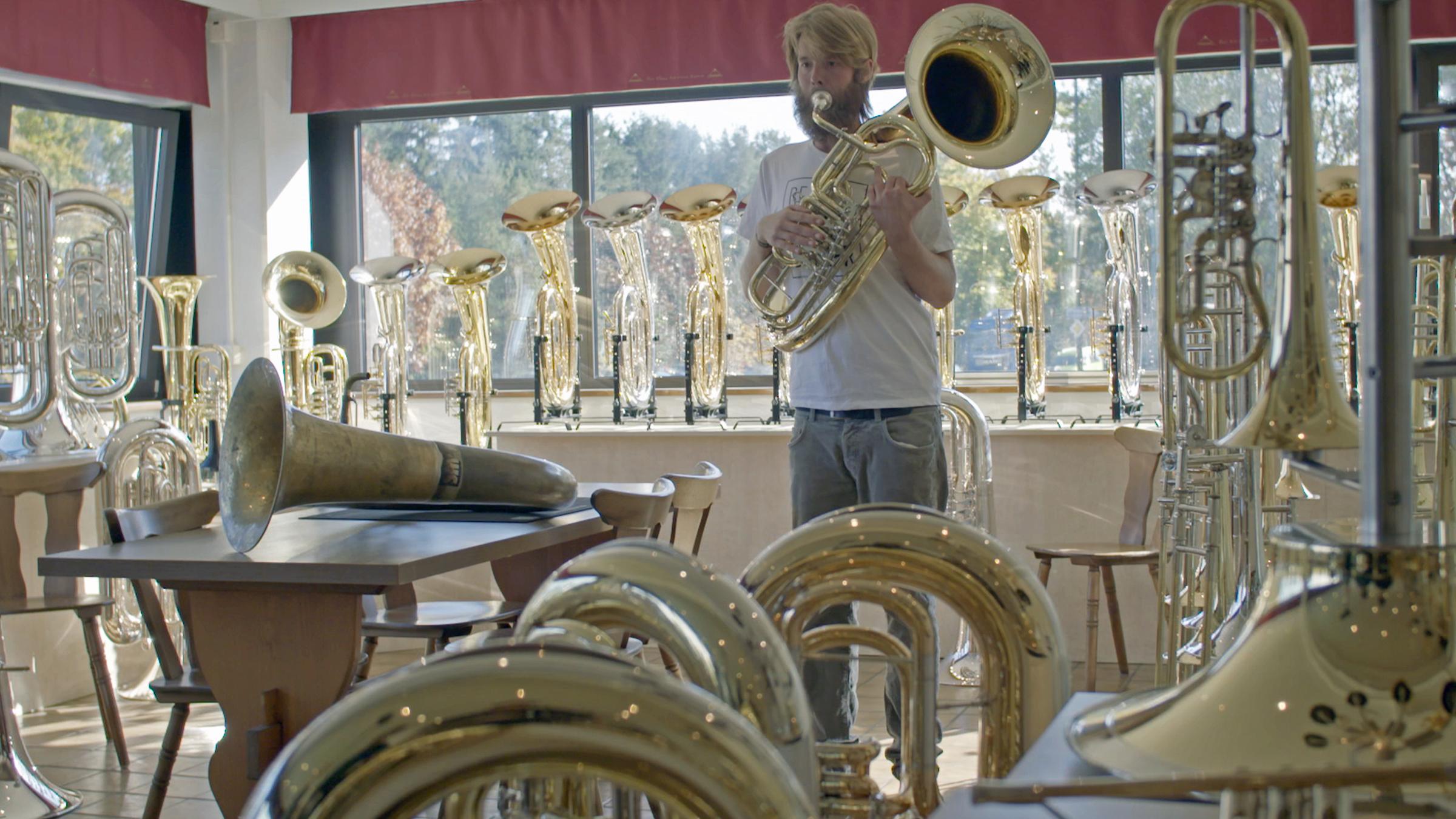 "plan b: Faire Arbeit - gerechter Lohn: Wie der Job uns glücklich machen kann": Stefan Huber steht in einem Raum voller Blasmusikinstrumente und spielt auf seiner Tuba.