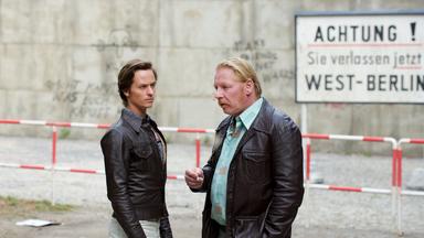 "Der gleiche Himmel (Teil 2)": Lars Weber (Tom Schilling) und Ralf Müller (Ben Becker) stehen unweit vom Sperrbezirk und unterhalten sich. Im Hintergrund ist die Berliner Mauer und ein Schild mit der Aufschrift "Achtung, Sie verlassen jetzt West-Berlin" zu sehen .