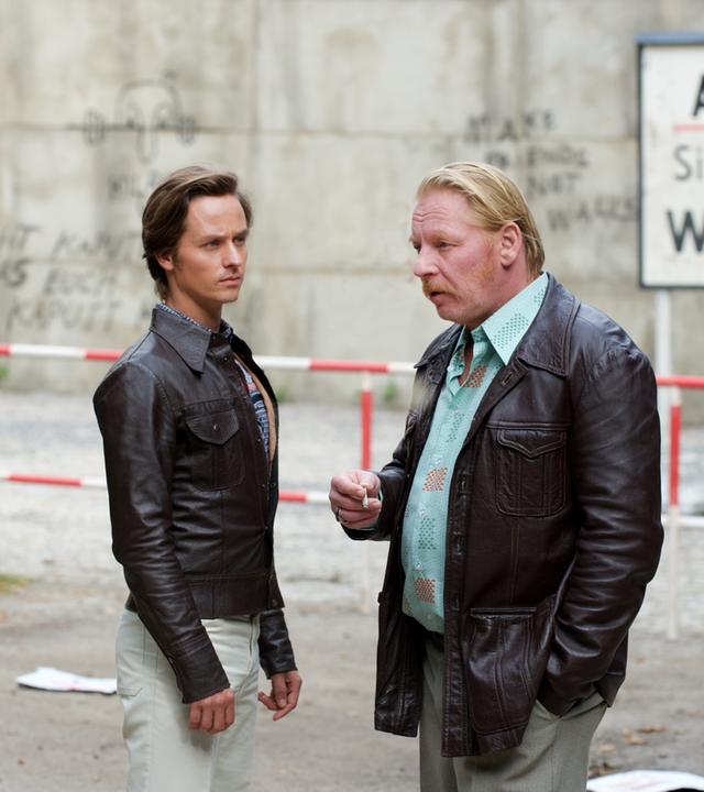 "Der gleiche Himmel (Teil 2)": Lars Weber (Tom Schilling) und Ralf Müller (Ben Becker) stehen unweit vom Sperrbezirk und unterhalten sich. Im Hintergrund ist die Berliner Mauer und ein Schild mit der Aufschrift "Achtung, Sie verlassen jetzt West-Berlin" zu sehen .