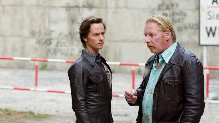 "Der gleiche Himmel (Teil 2)": Lars Weber (Tom Schilling) und Ralf Müller (Ben Becker) stehen unweit vom Sperrbezirk und unterhalten sich. Im Hintergrund ist die Berliner Mauer und ein Schild mit der Aufschrift "Achtung, Sie verlassen jetzt West-Berlin" zu sehen .