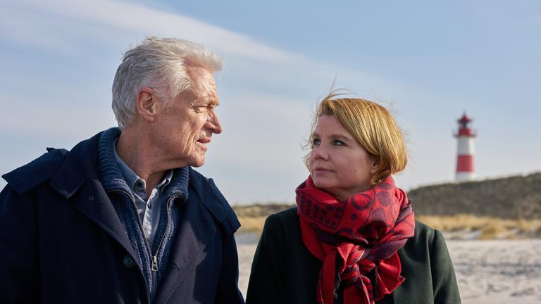 "Nord Nord Mord - Clüver und die tödliche Affäre": Bernadette Kipling (Annette Frier) und Theo Clüver (Robert Atzorn) gehen an einem sonnigen Herbsttag am Strand spazieren. Im Hintergrund rechts ist ein  Leuchtturm zu sehen.