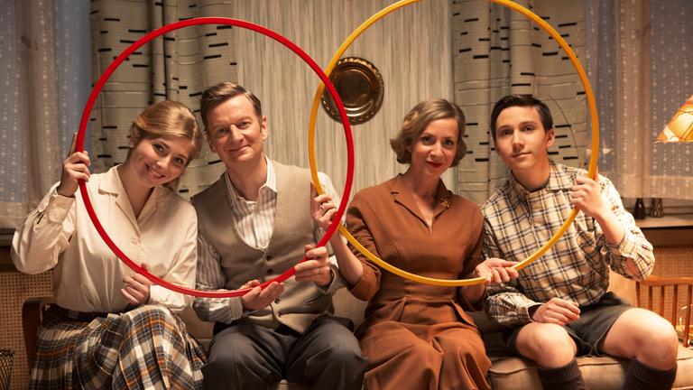 "ZDFzeit: Das Jahrhunderthaus - Essen, Trinken und Sport": Tochter Lena (Lilian Prent), Vater Thomas (Michael Kessler), Mutter Anna (Ruth Blauert) und Sohn Alex (Benedict Jacob) sitzen im Wohnzimmer auf einem Sofa. Lena und Thomas halten gemeinsam einen Hula-Hoop-Reifen in der Hand, ebenso wie Anna und Alex.