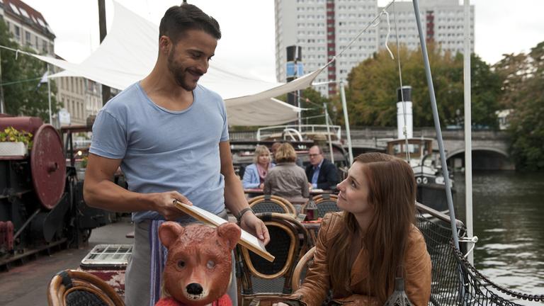 "Lotta & der Ernst des Lebens": Lotta (Josefine Preuß) und Lilo (Matilda Jork) mit Bärchenmaske sitzen am Tisch, Junis (Kostja Ullmann) bringt die Speisekarte. Im Hintergrund: Häuser, Menschen und Wasser.