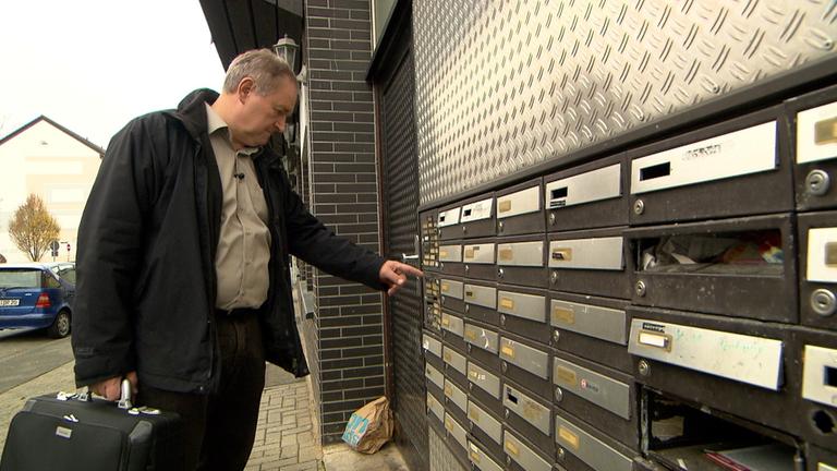 "In der Schuldenfalle - Zwischen Kontopfändung und Gerichtsvollzieher": Gerichtsvollzieher Michael Peller steht am Hauseingang eines Mehrfamilienhauses und blickt suchend, mit ausgestrecktem Finger, auf die einzelnen Klingelschilder.