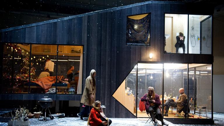"Drei Schwestern": Das Foto zeigt eine Holzhütte im Schnee. Drei Zimmer sind erleuchtet. Links im Bild ist das Wohnzimmer zu sehen, das weihnachtlich geschmückt ist. Auf dem Sofa liegt ein Mann und schläft. Er ist mit einer Wolldecke zugedeckt und schläft. Es sind noch 2 weitere Männer und eine Frau im Raum. Sie sehen sich aber nicht an: einer spielt Klavier, die Frau sitzt auf dem Sofa und der andere Mann hat den Rücken dem Betrachter zugewandt. Rechts kann man in das erleuchtet Fenster der Küche sehen, die nicht aufgeräumt ist. Dort sitzt ein Mann am Tisch und liest. Über der Küche ist das Bad hell erleuchtet. Dort sitzt auf dem Klodeckel eine Frau in schwarz, die ihr Gesicht in den Händen verbirgt und weint. Auf der Terrasse im Vordergrund des Bildes liegt Schnee. Dort halten sich zwei Männer und eine Frau auf. Die Frau sitzt in einem roten Mantel und dickem Schal auf der Terrassenstufe und scheint sich mit dem Mann rechts im Bild zu unterhalten. Der grauhaarige Mann sitzt auf einem Stuhl. Er ist mit einer Jeans und einem lila Wollmantel bekleidet. In einer Hand hält er eine Brille.