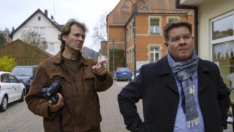 "37° - Der Bürgermeister-Macher": Abberger steht mit seinem Kandidaten Zeilmeier im Ort Ispringen. Abberger hält rechts eine Kamera in der Hand und mit der linken Hand zeigt er Zeilmeier, wo er ihn fotografieren will.