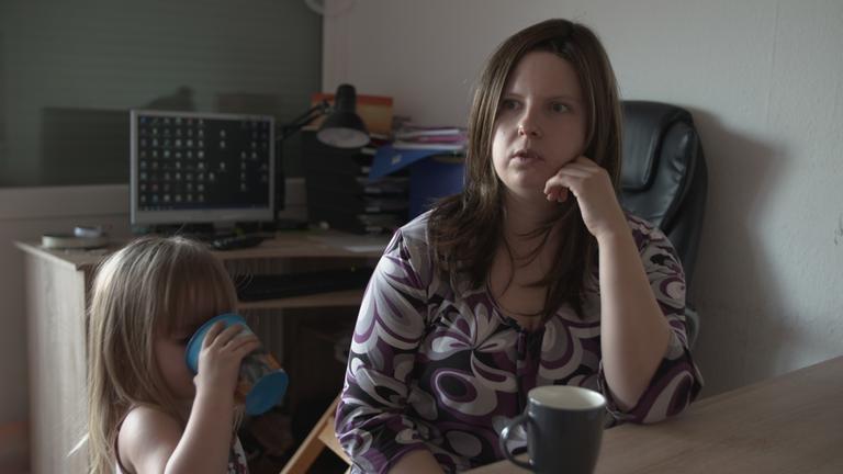 "Vergessen im Westen" (3sat): In der Mitte des Bildes sitzt eine Frau am Tisch. Neben ihr steht ihre kleine Tochter und trinkt aus einem Becher. Im Hintergrund ein Computermonitor  auf einem Schreibtisch