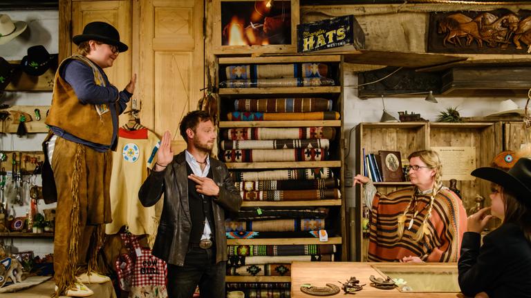 "Winnetous Sohn": Auf der linken Seite steht Max (Lorenzo Germeno) im Indianerkostüm auf der Theke eines Ladens für Wild-West-Ausstattung. Sein Vater Torsten (Christoph Letkowski) steht davor. Beide zeigen das indianische Friedens- und Freundschaftszeichen. Morten (Tristan Göbel) und eine Verkäuferin beobachten diese Gesten skeptisch.