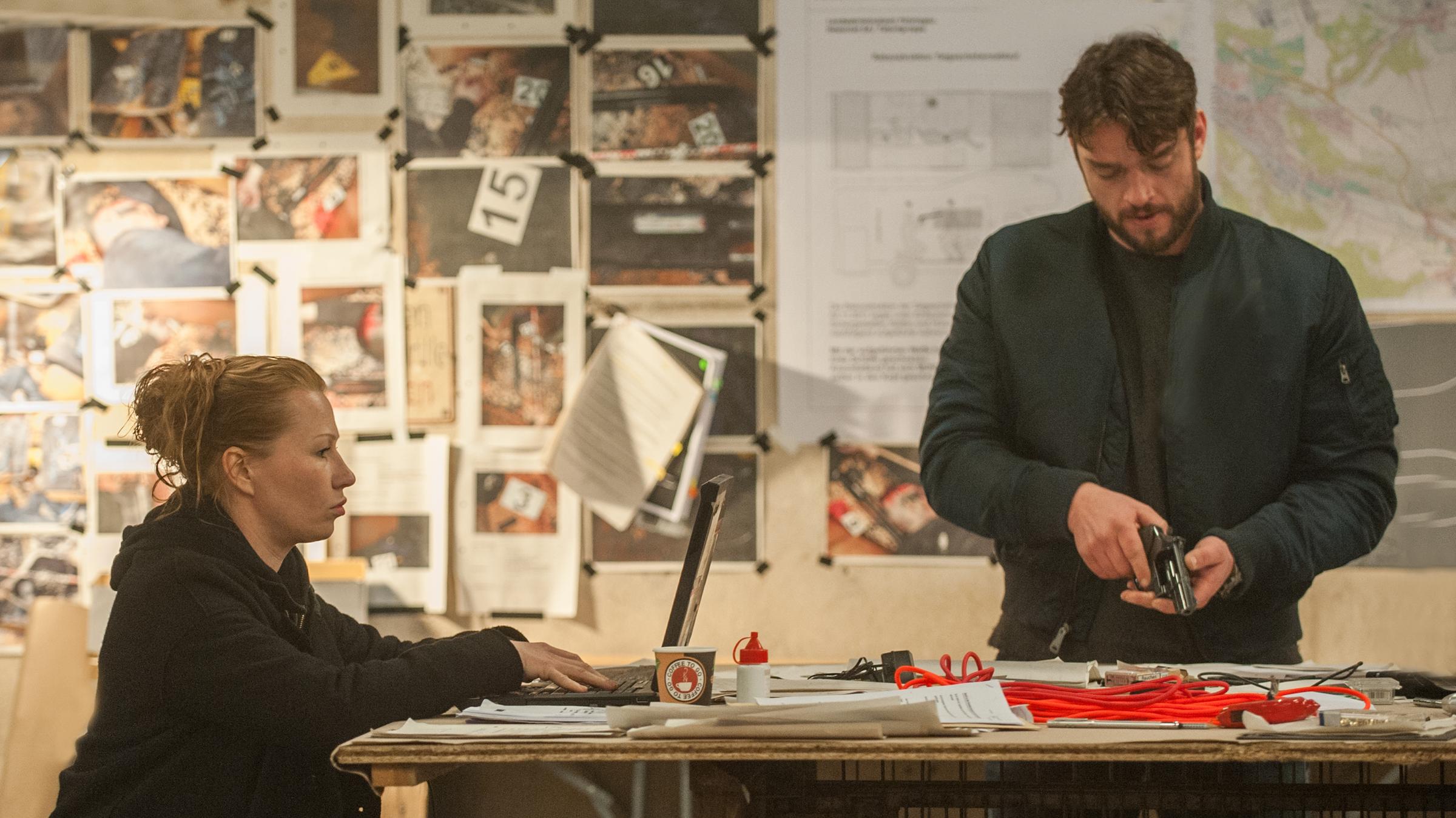 "Dengler - Die schützende Hand": Olga (Birgit Minichmayr) sitzt an einem improvisierten Schreibtisch und arbeitet an ihrem Laptop. Dengler (Ronald Zehrfeld) steht neben ihr und lädt seine Pistole. An der Wand im Hintergrund sind zahlreiche Tatortfotos und andere Schriftstücke angepinnt.