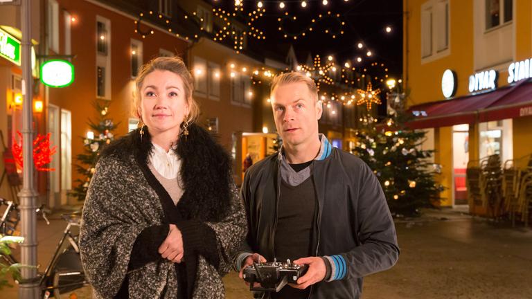 "Der mit dem Schlag": Melanie Merk (Lisa Maria Potthoff) und Felix Grünler (Hinnerk Schönemann) stehen nebeneinander auf einer weihnachtlich beleuchteten Straße. Felix hat eine Fernbedienung in seinen Händen. Er und Melanie schauen erwartungsvoll nach vorne.