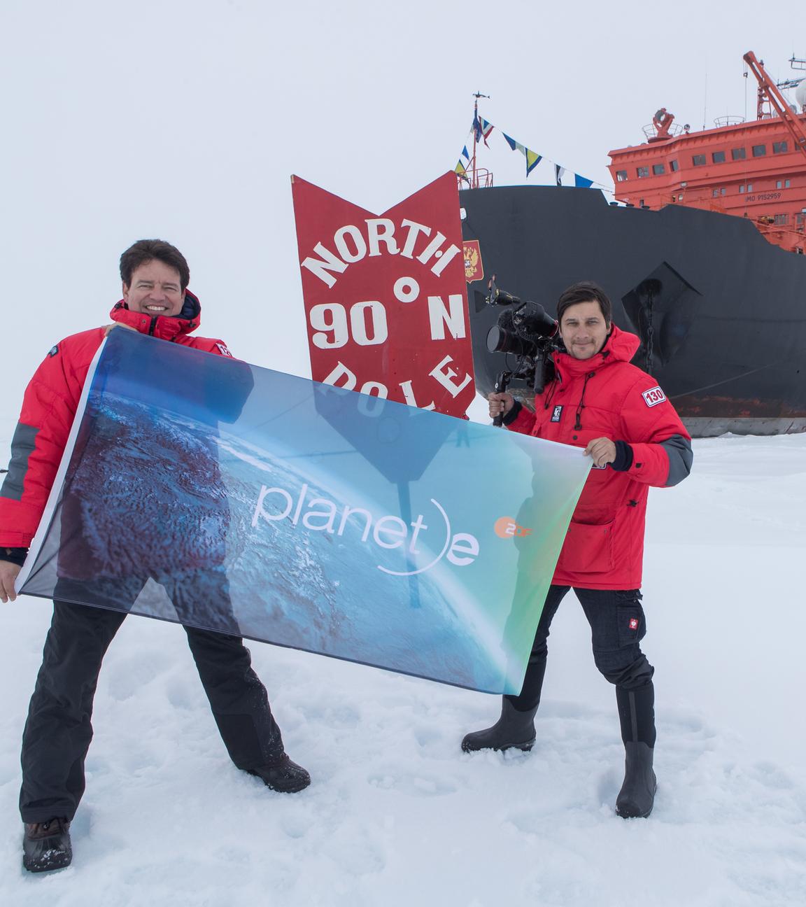 "planet e.: Kampf um die Arktis": Autor Andreas Ewels steht mit Kameramann Aris Bibudis im Eis. Die beiden halten eine "planet e."-Fahne hoch. Hinter ihnen ist das Schiff "50 Years of Victory" zu sehen.