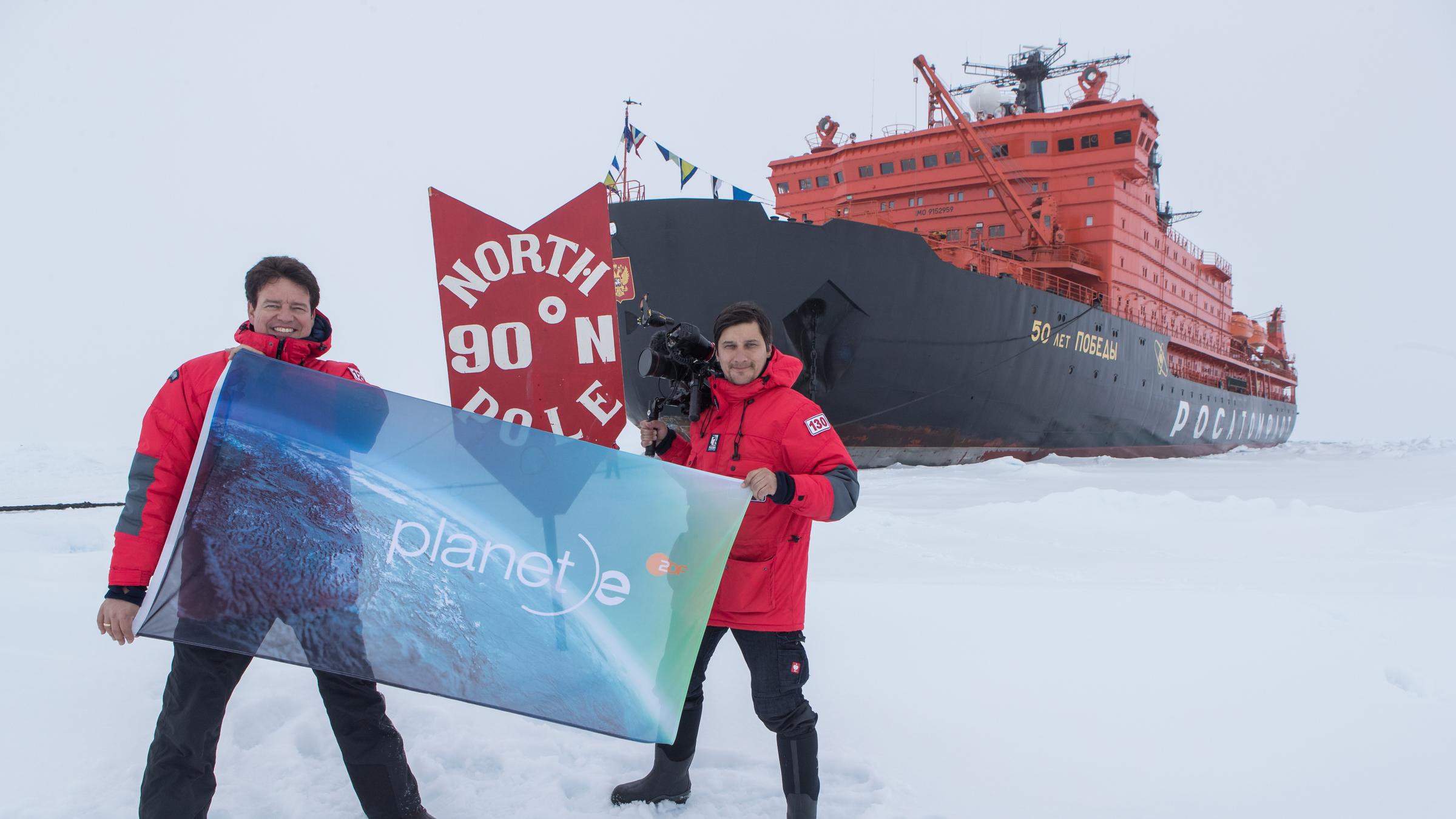 "planet e.: Kampf um die Arktis": Autor Andreas Ewels steht mit Kameramann Aris Bibudis im Eis. Die beiden halten eine "planet e."-Fahne hoch. Hinter ihnen ist das Schiff "50 Years of Victory" zu sehen.