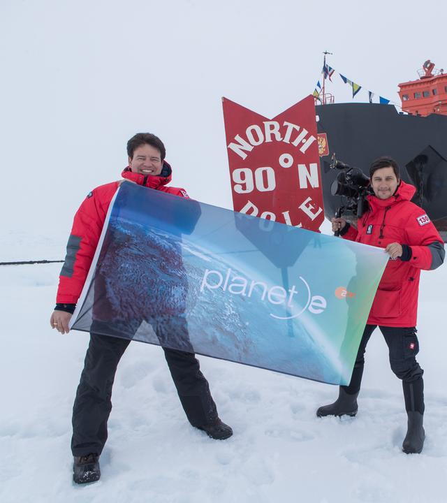 "planet e.: Kampf um die Arktis": Autor Andreas Ewels steht mit Kameramann Aris Bibudis im Eis. Die beiden halten eine "planet e."-Fahne hoch. Hinter ihnen ist das Schiff "50 Years of Victory" zu sehen.