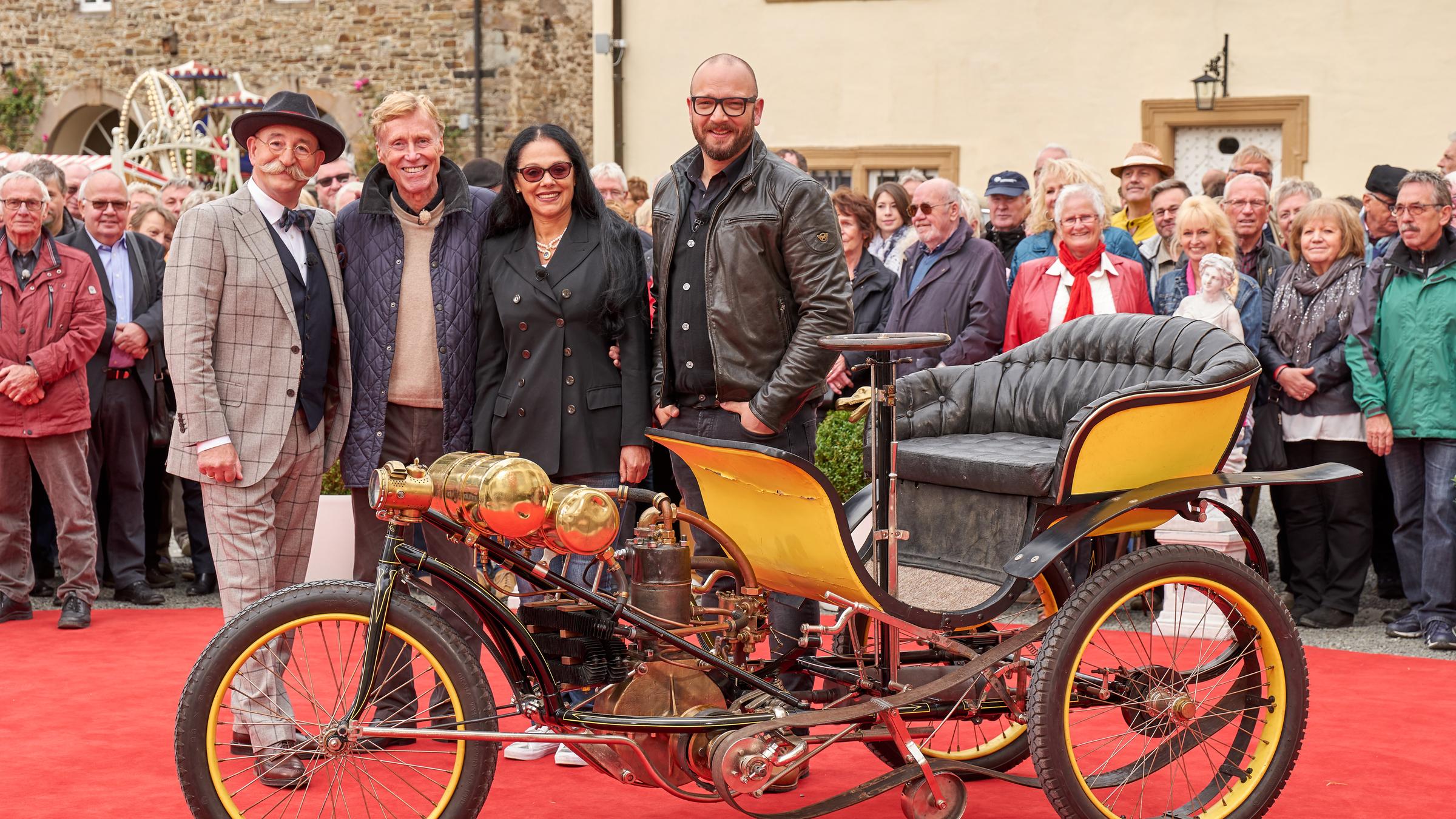 "Bares für Rares": Moderator Horst Lichter steht mit zwei Kandidaten und dem Experten Sven Deutschmanek vor einer alten Fahrradkutsche. Im Hintergrund steht das Publikum.