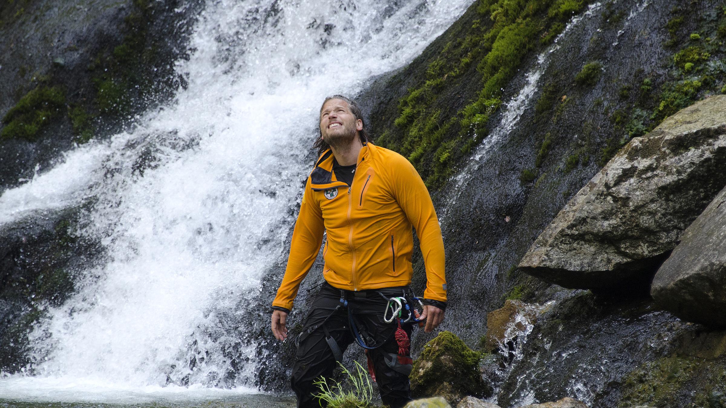 "Die Bergretter: Am Abgrund": Markus (Sebastian Ströbel) steht am Ende eines Wasserfalls, den Blick fragend nach oben gerichtet. Er trägt einen Rettungsgurt, an dem verschiedene Karabiner befestigt sind.