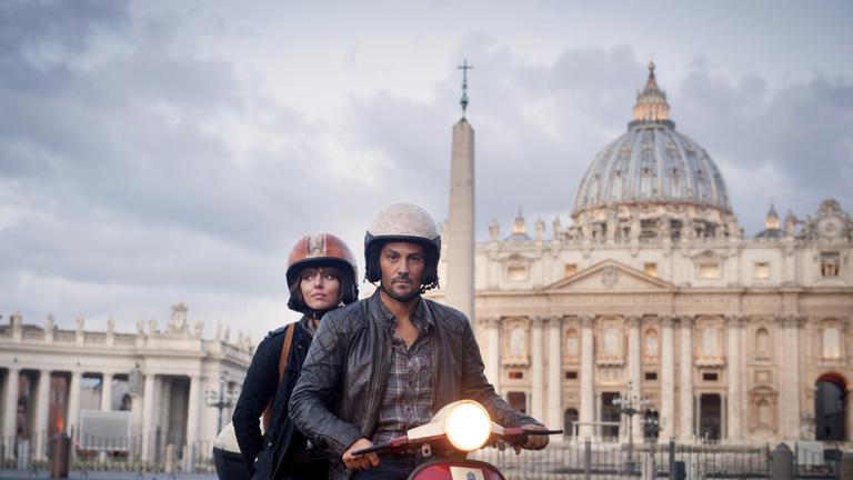 "Hooten & the Lady - Alle Wege führen nach Rom": Alex (Ophelia Lovibond) und Hooten (Michael Landes) sitzen auf einer Vespa auf dem Petersplatz in Rom.