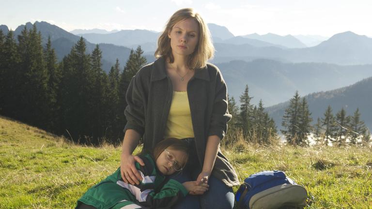 "Eine unerhörte Frau": Hanni (Rosalie Thomass) sitzt auf einer Wiese. Ihrer Tochter Magdalena (Romy Butz) liegt auf ihrem Schoß. Beide schauen ernst.