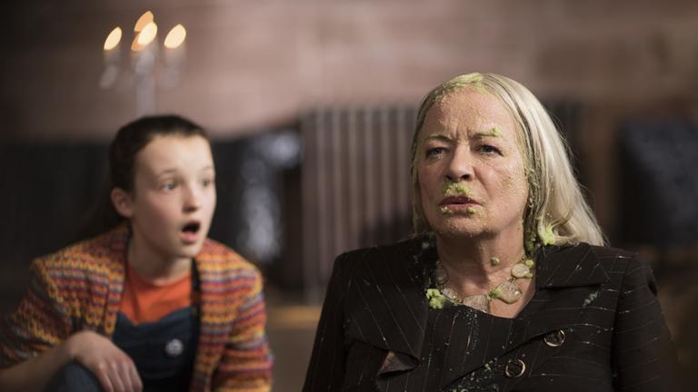 "Eine lausige Hexe - Die Aufnahmeprüfung (2)": Mildred Hoppelt (Bella Ramsey) hat Augen und Mund weit aufgerissen, sie kniet mit einem Knie auf dem Boden und blickt zu Frau Agatha Graustein (Clare Higgins), die neben ihr auf dem Boden sitzt. Agatha blickt starr nach vorne und ist über und über mit grüner Suppe bekleckert.