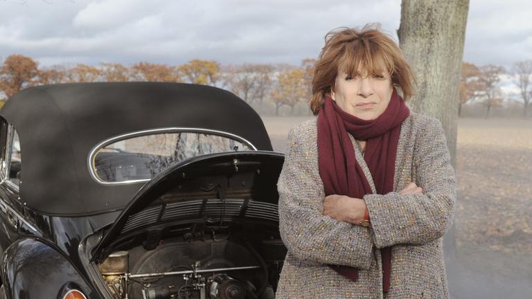 "Bella Block - Stille Wasser": Bella Block (Hannelore Hoger) steht in einer Feldlandschaft mit verschränkten Armen vor der geöffneten Heckklappe ihres VWs und wartet auf den Abschleppdienst.