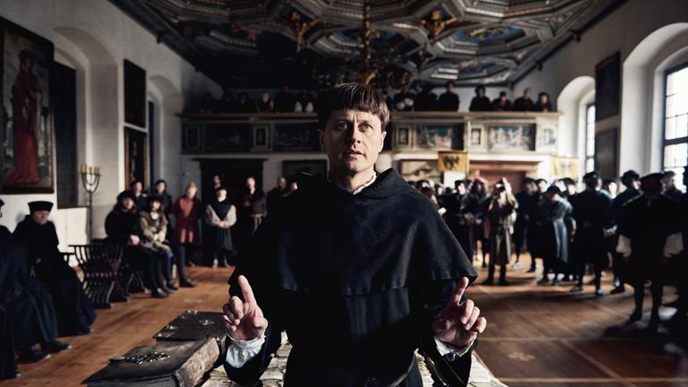 „Das Luther-Tribunal – Zehn Tage im April“: Martin Luther (Roman Knižka) steht bei der Hauptverhandlung vor dem Tisch, auf dem seine Schriften ausgelegt sind