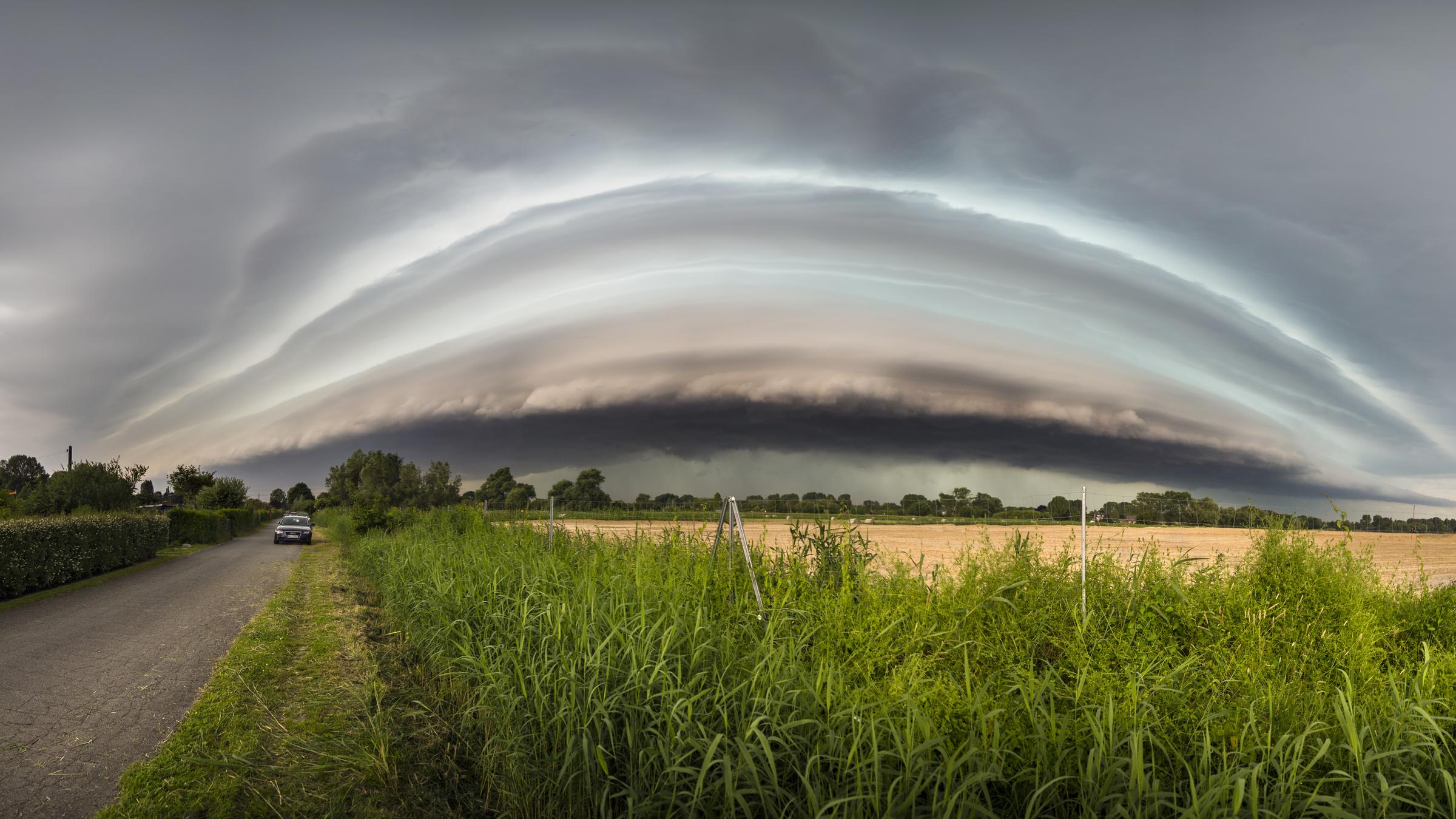 "ZDF.reportage - Jagd aufs Unwetter": Am Himmel braut sich ein Gewitter zusammen. Ein Auto fährt an einem Feld entlang.