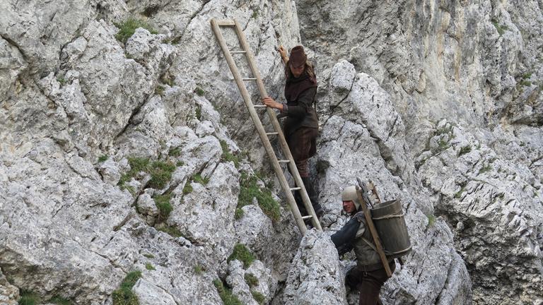 "Die Alpen - Eine große Geschichte". Szene: Zwei Darsteller versuchen mit einer Leiter einen Berg zu besteigen.  Im Hintergrund Felswand.