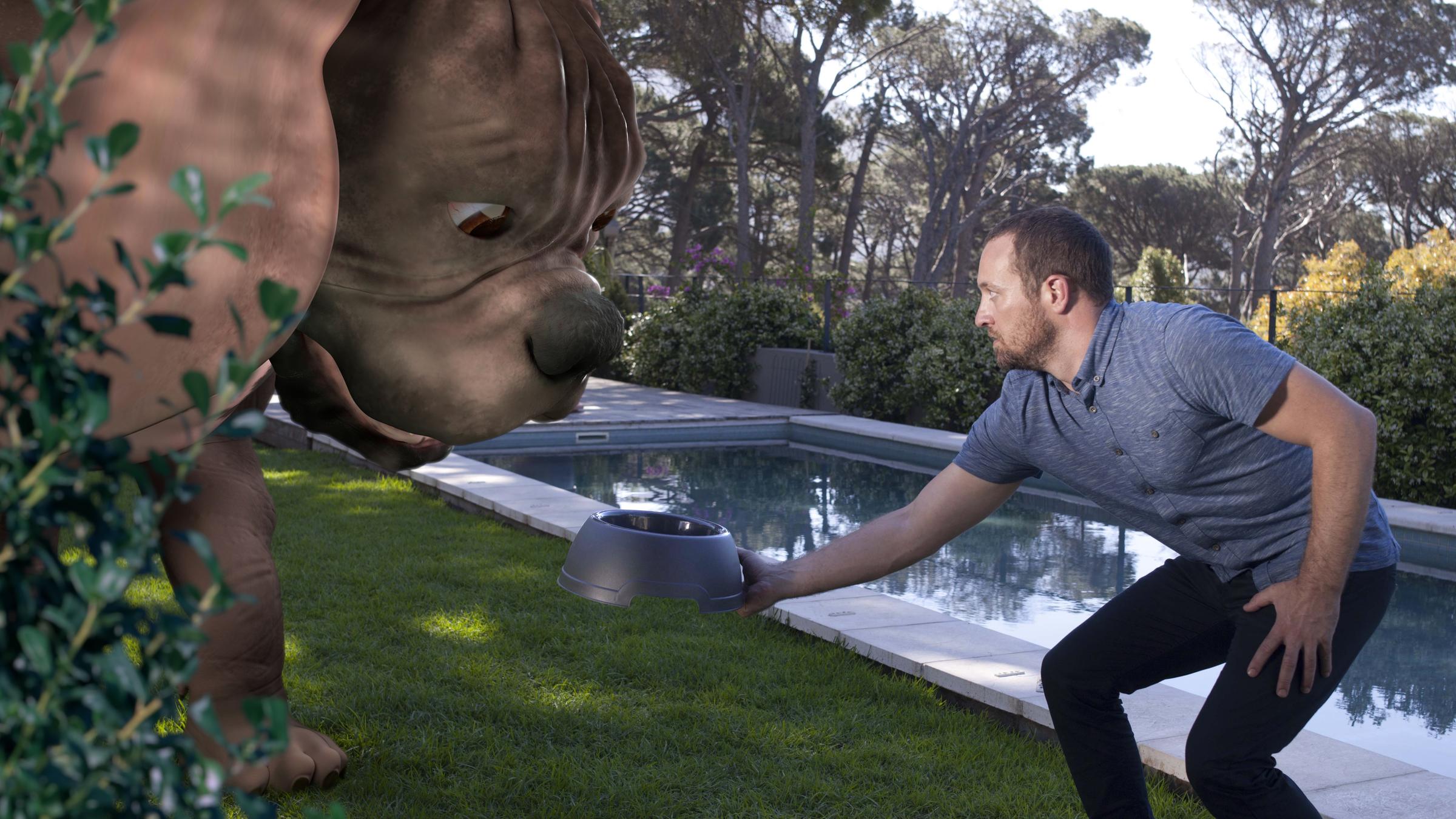 "Terra X: Auf die Größe kommt es an - Riesen": Ein Mann hält einem Riesenhund aus Plastik einen Fressnapf hin. Er sieht sehr ängstlich aus. Im Hintergrund ist ein Garten mit einem Schwimmbecken zu sehen.