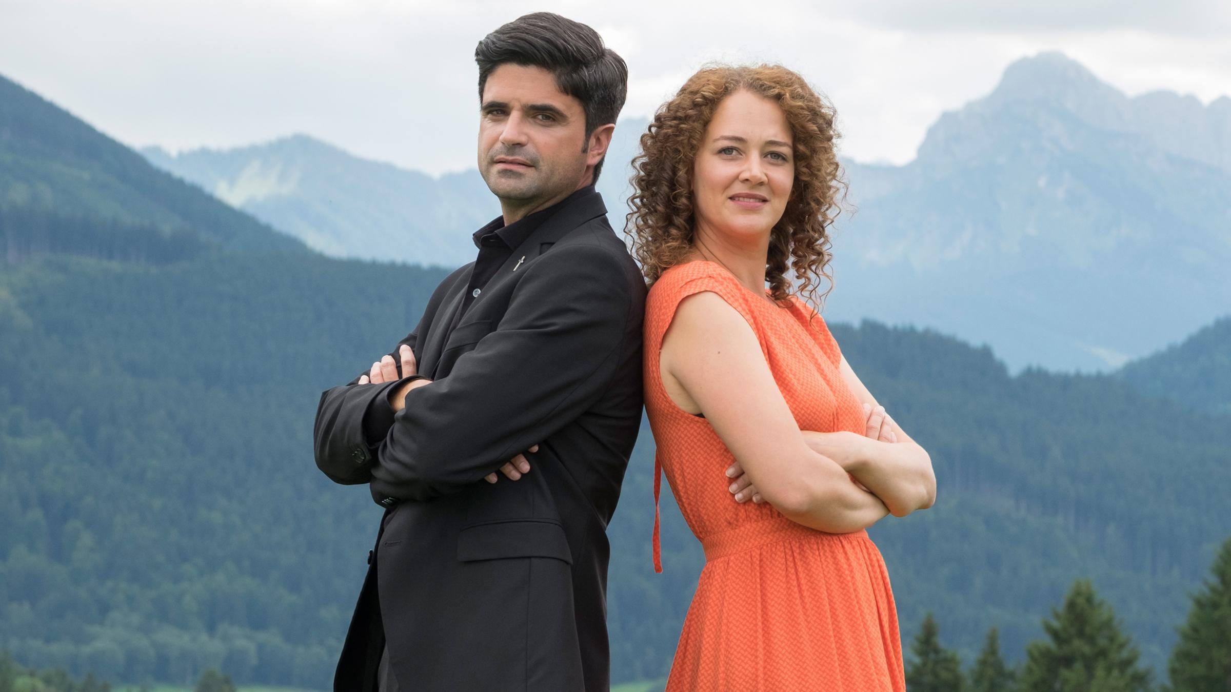"Tonio und Julia - Kneifen gilt nicht": Mit dem Rücken aneinandergelehnt und nach vorne in die Kamera blickend stehen Pfarrer Tonio Niederegger (Maximilian Grill) und Familientherapeutin Julia Schindel (Oona Devi Liebich) auf einer Wiese. Im Bildhintergrund Bäume und Berge.