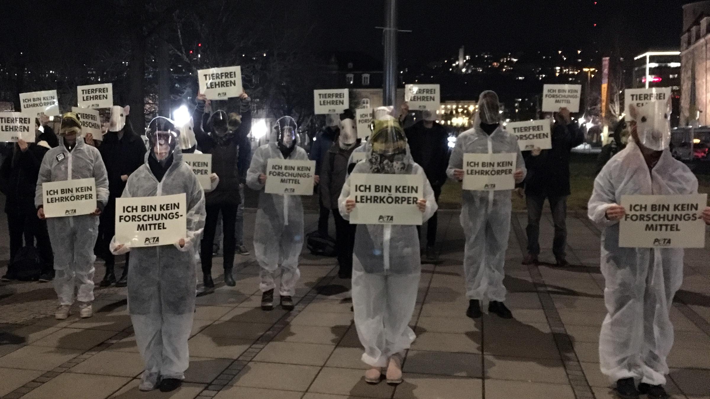 "planet e.: Tierversuche auf dem Prüfstand": Eine Gruppe Menschen in weißen Schutzanzügen und mit Tiermasken über dem Gesicht steht im Dunkeln auf einem Platz. Alle halten Protestplakate in ihren Händen, die mit "PETA" unterzeichnet sind. Auch Personen ohne Schutzanzüge sind dabei.