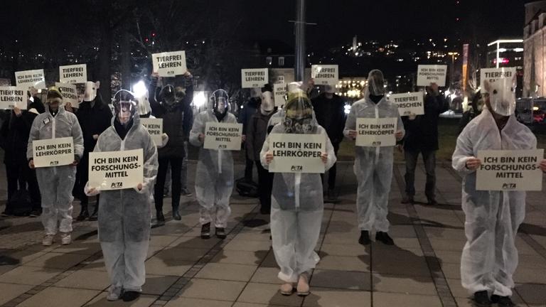 "planet e.: Tierversuche auf dem Prüfstand": Eine Gruppe Menschen in weißen Schutzanzügen und mit Tiermasken über dem Gesicht steht im Dunkeln auf einem Platz. Alle halten Protestplakate in ihren Händen, die mit "PETA" unterzeichnet sind. Auch Personen ohne Schutzanzüge sind dabei.
