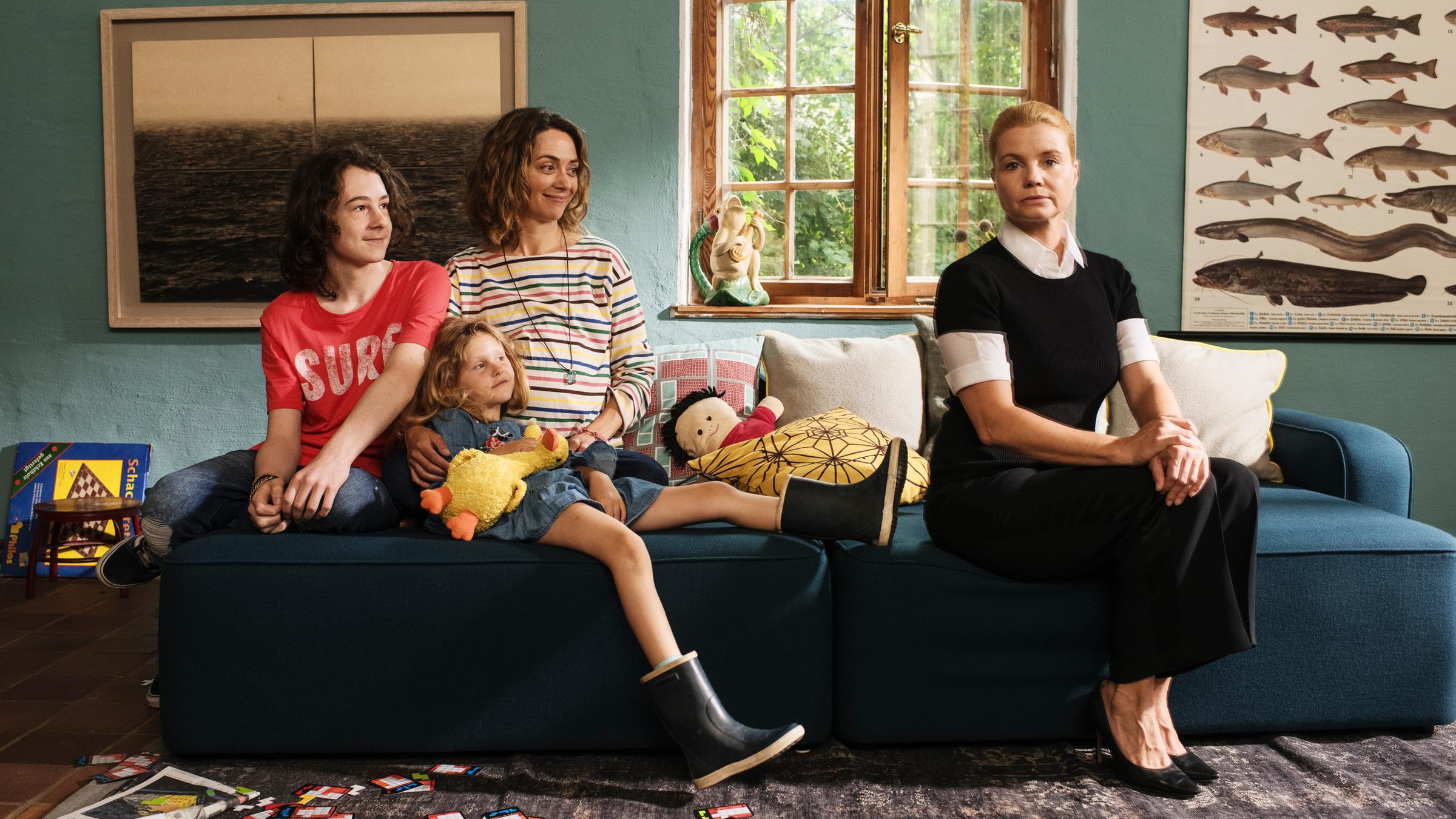 "Ella Schön - Die Inselbegabung": Ben Kieper (Maximilian Ehrenreich), Klara Kieper (Zora Müller), Christina Kieper (Julia Richter) und Ella Schön (Annette Frier) sitzen gemeinsam auf einem Sofa im Wohnzimmer. Ella sitzt sehr aufrecht und schaut starr nach vorne, die Kiepers haben es sich auf der Couch gemütlich gemacht und beobachten Ella amüsiert.